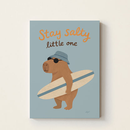 Hand-drawn capybara wearing sunglasses and a blue bucket hat while holding a surfboard beneath the words "Stay Salty Little One", whimsical coastal wall art for nurseries, bedrooms, and playrooms.